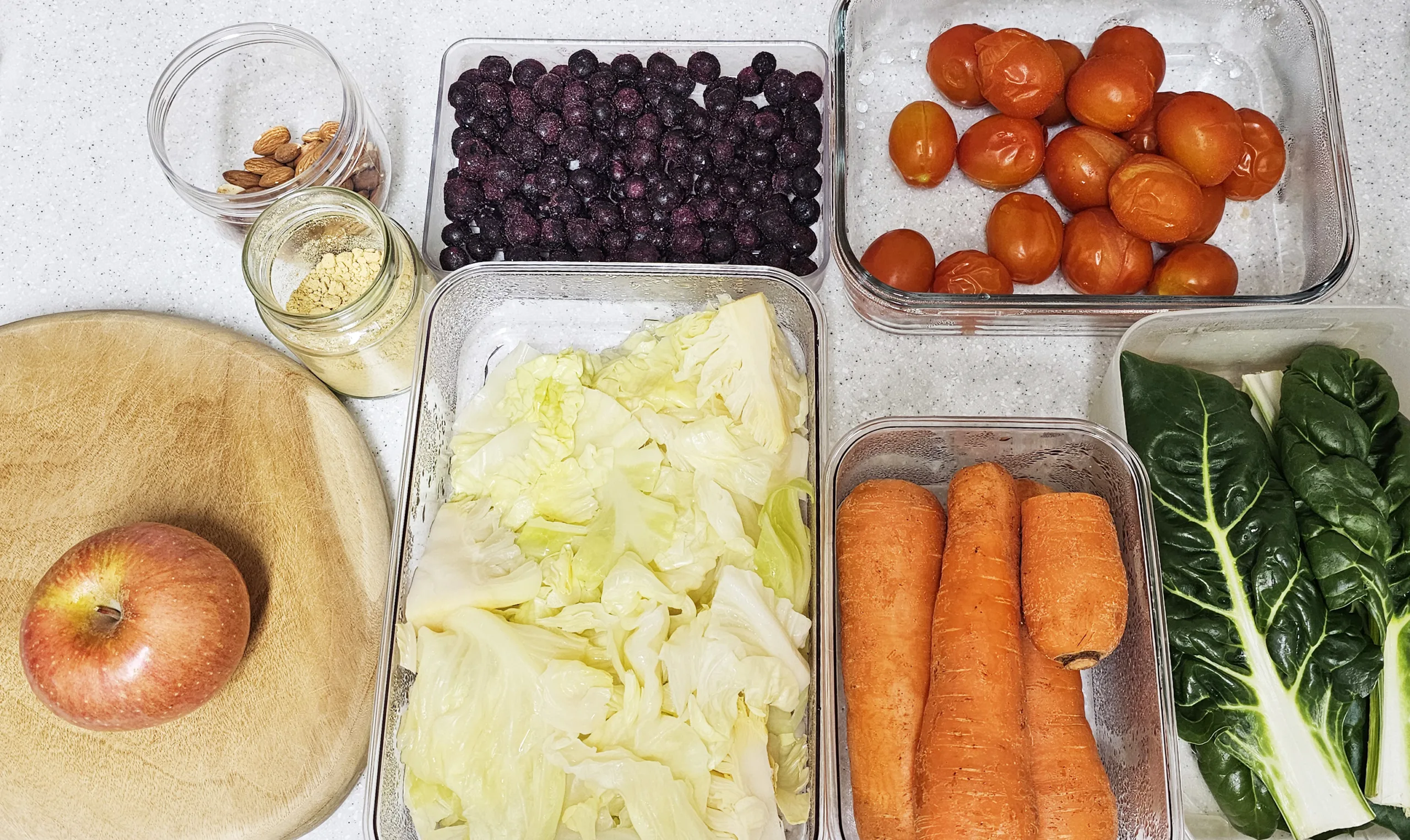 해독주스 만들기 재료들이 테이블위에 준비되어 있다. 냉동블루베리, 양배추, 당근, 근대, 사과, 대추방울토마토, 아몬드, 생강가루가 밀폐통에 담겨있다.